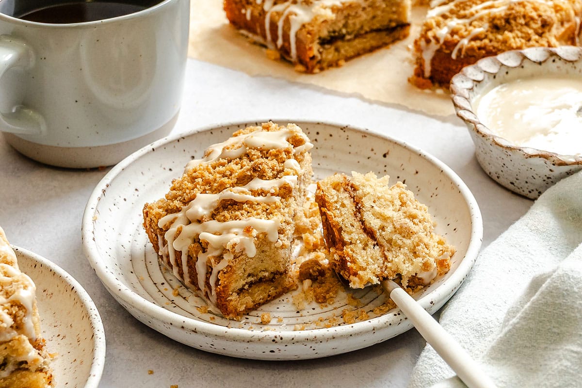 A plate with a slice of crumb cake drizzled with icing, a fork cutting into it, a cup of coffee, and bowls of icing sit on a light surface.
