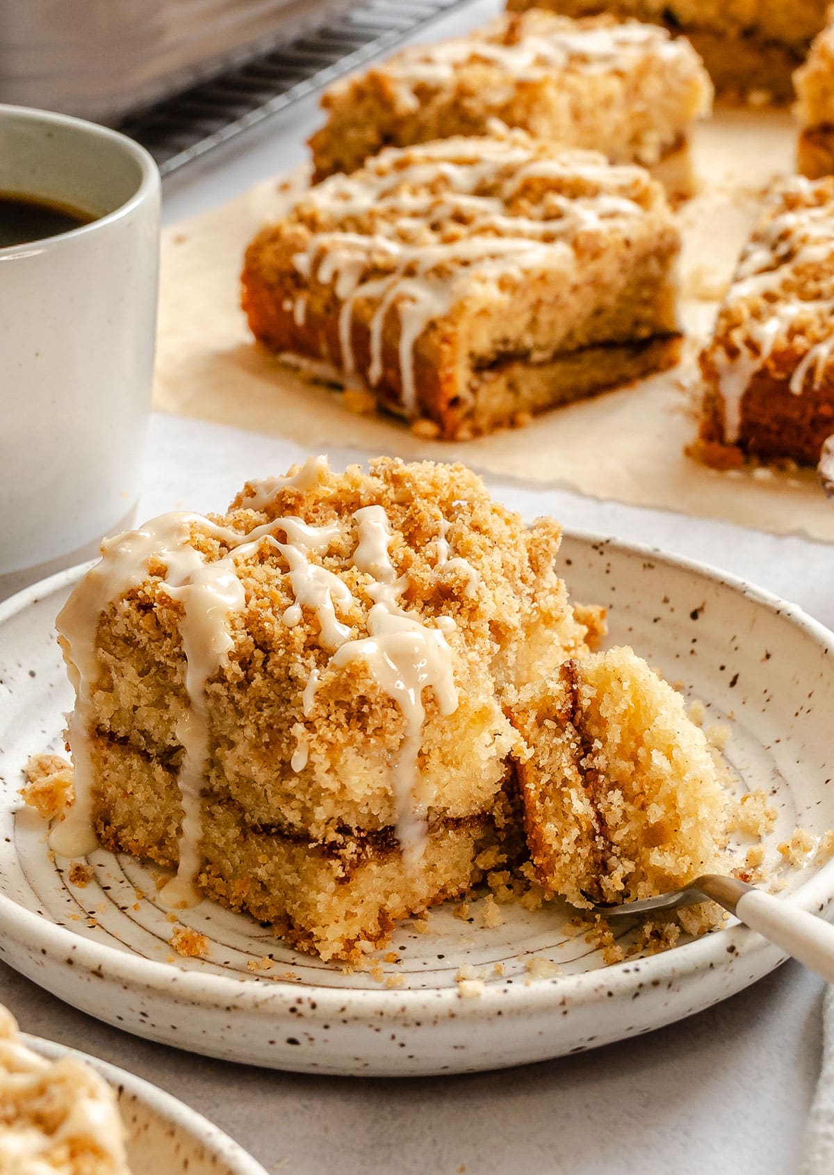 A plate with two stacked pieces of crumb-topped vegan coffee cake drizzled with icing, with a fork taking a bite and a cup of coffee in the background.