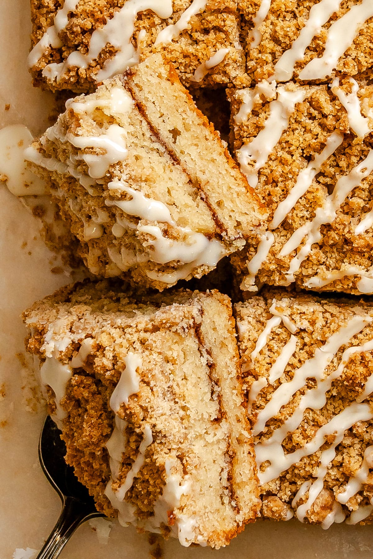 Sliced coffee cake with crumb topping and white icing drizzle, arranged on parchment paper with a serving utensil.