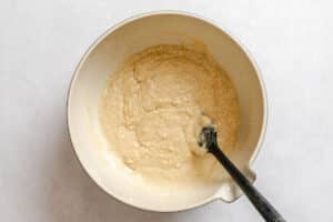 A white mixing bowl with light-colored batter and a black spatula on a white surface.