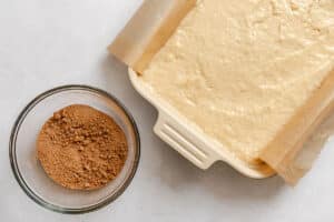A baking dish lined with parchment paper is filled with cake batter next to a glass bowl containing crumbly brown topping, all on a light surface.
