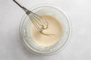 A metal whisk is resting in a glass bowl filled with smooth, pale batter on a light gray surface.