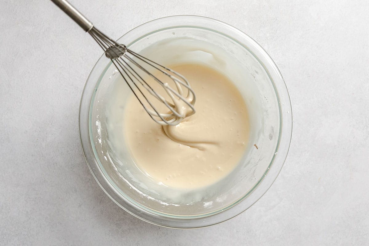 A metal whisk is resting in a glass bowl filled with smooth, pale batter on a light gray surface.