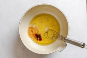 A mixing bowl with applesauce, oil, and vanilla extract, with a metal whisk resting inside, on a light-colored surface.