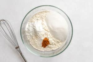 A glass bowl containing flour, sugar, baking powder, and cinnamon on a white surface next to a metal whisk.