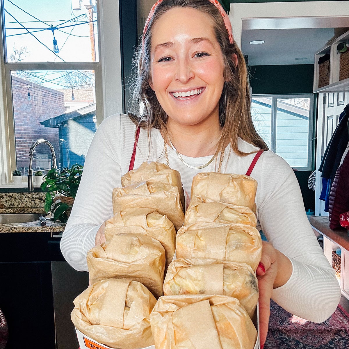 A woman smiling and holding a container filled with several vegetarian breakfast sandwiches wrapped in brown paper in a kitchen.