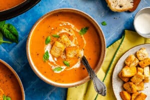 A bowl of tomato soup topped with croutons, cream, and basil leaves sits on a blue surface next to a plate of croutons, a slice of bread, and a napkin.