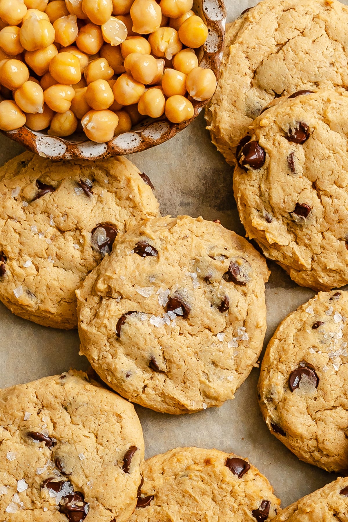 Chocolate chip chickpea cookies arranged on a surface next to a bowl filled with chickpeas.