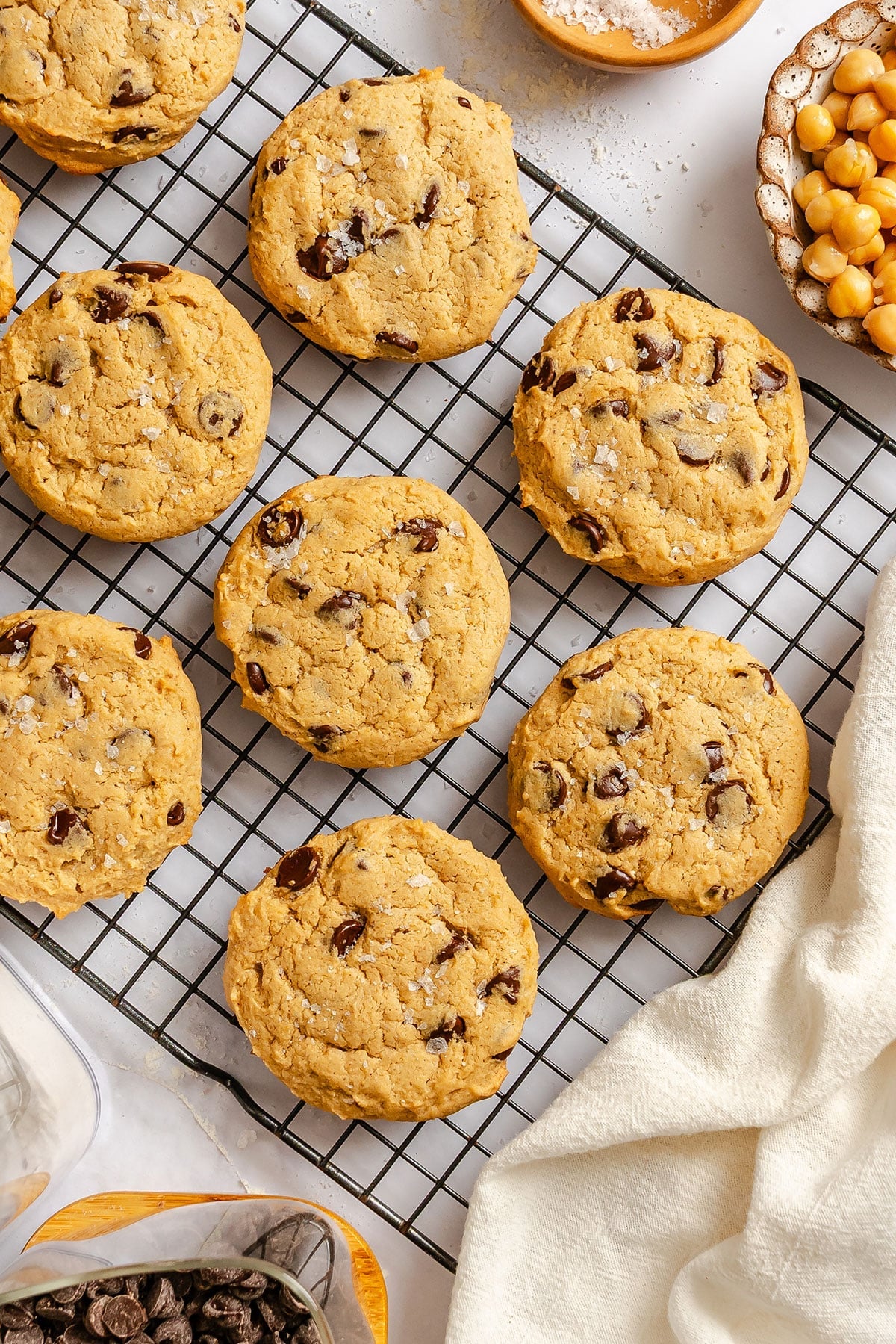 Chocolate chip chickpea cookies cooling on a wire rack, surrounded by a bowl of chickpeas, a bowl of salt, chocolate chips, and a white cloth.