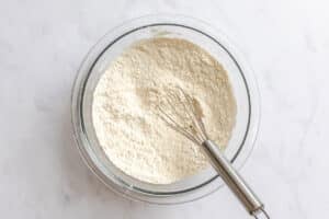 A glass mixing bowl filled with flour and a metal whisk resting inside, placed on a white countertop.