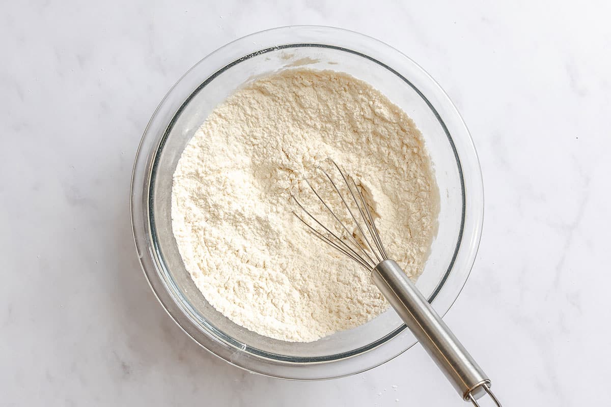 A glass mixing bowl filled with flour and a metal whisk resting inside, placed on a white countertop.