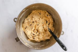A mixing bowl filled with chocolate chip cookie dough and a black spatula resting inside, on a white marble surface.