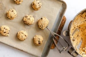 Scoops of chocolate chip cookie dough on a parchment-lined baking sheet, with a mixing bowl and cookie scoop nearby.