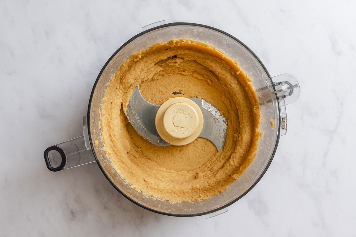 A food processor containing chickpea puree sits on a white marble surface.