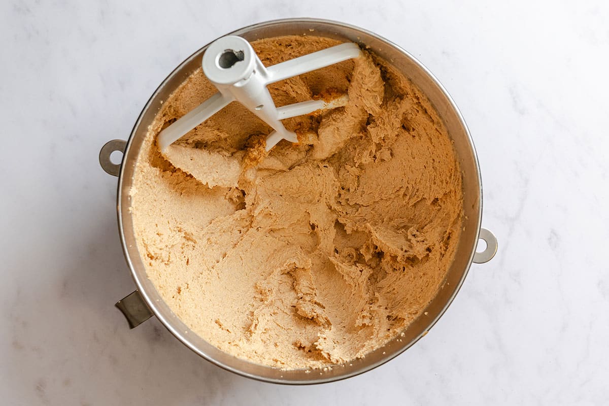 A mixing bowl with a paddle attachment contains creamed butter and sugar on a white surface.
