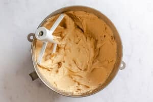A metal mixing bowl with a paddle attachment holds creamy, light brown cookie dough on a white marble surface.