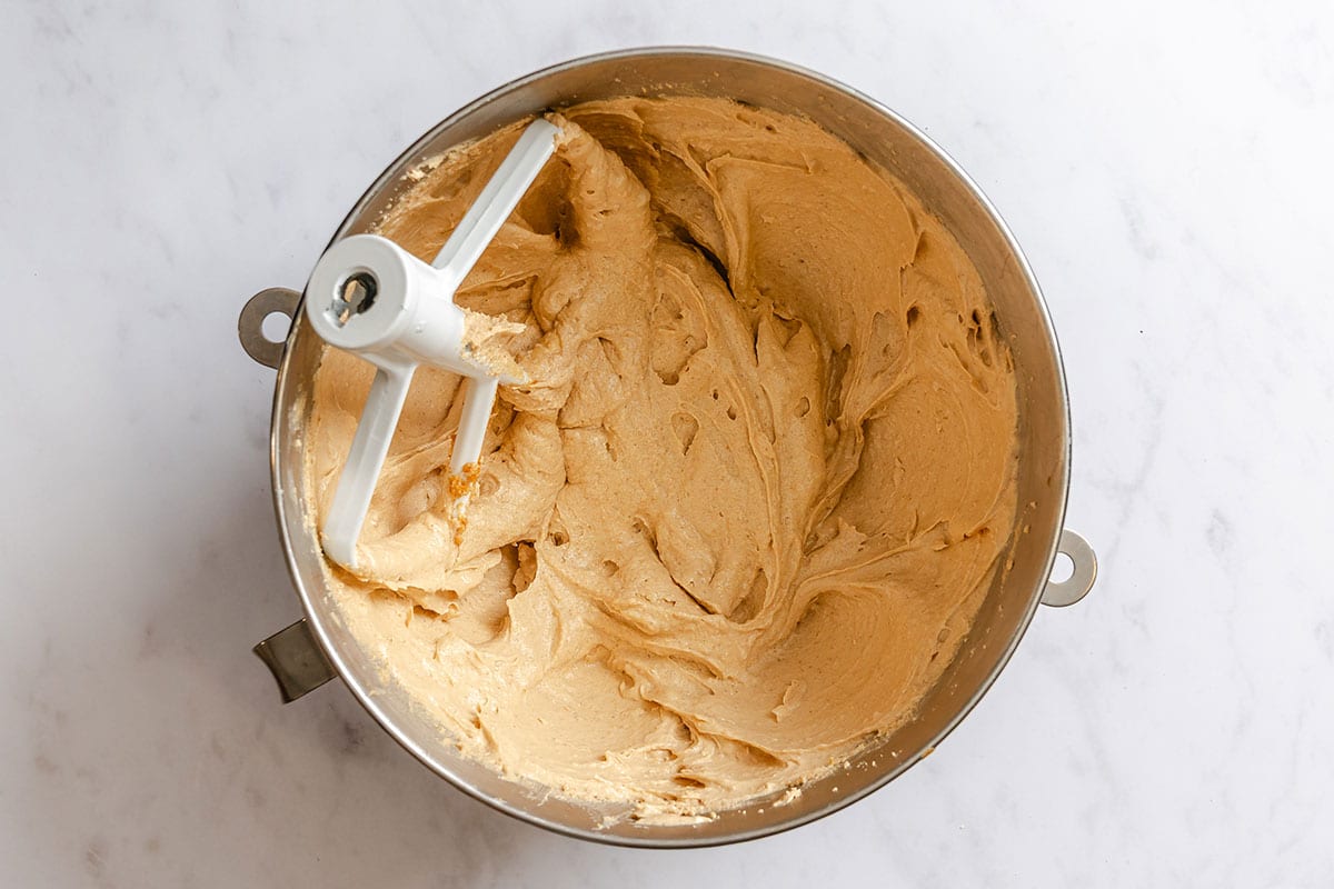 A metal mixing bowl with a paddle attachment holds creamy, light brown cookie dough on a white marble surface.