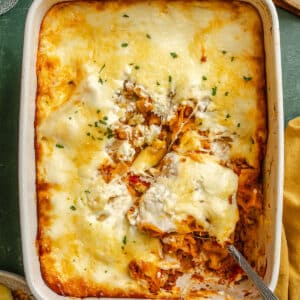 A baked pasta casserole topped with melted cheese sits in a white dish, with a serving missing and a spoon resting inside. Slices of bread and glasses are nearby.