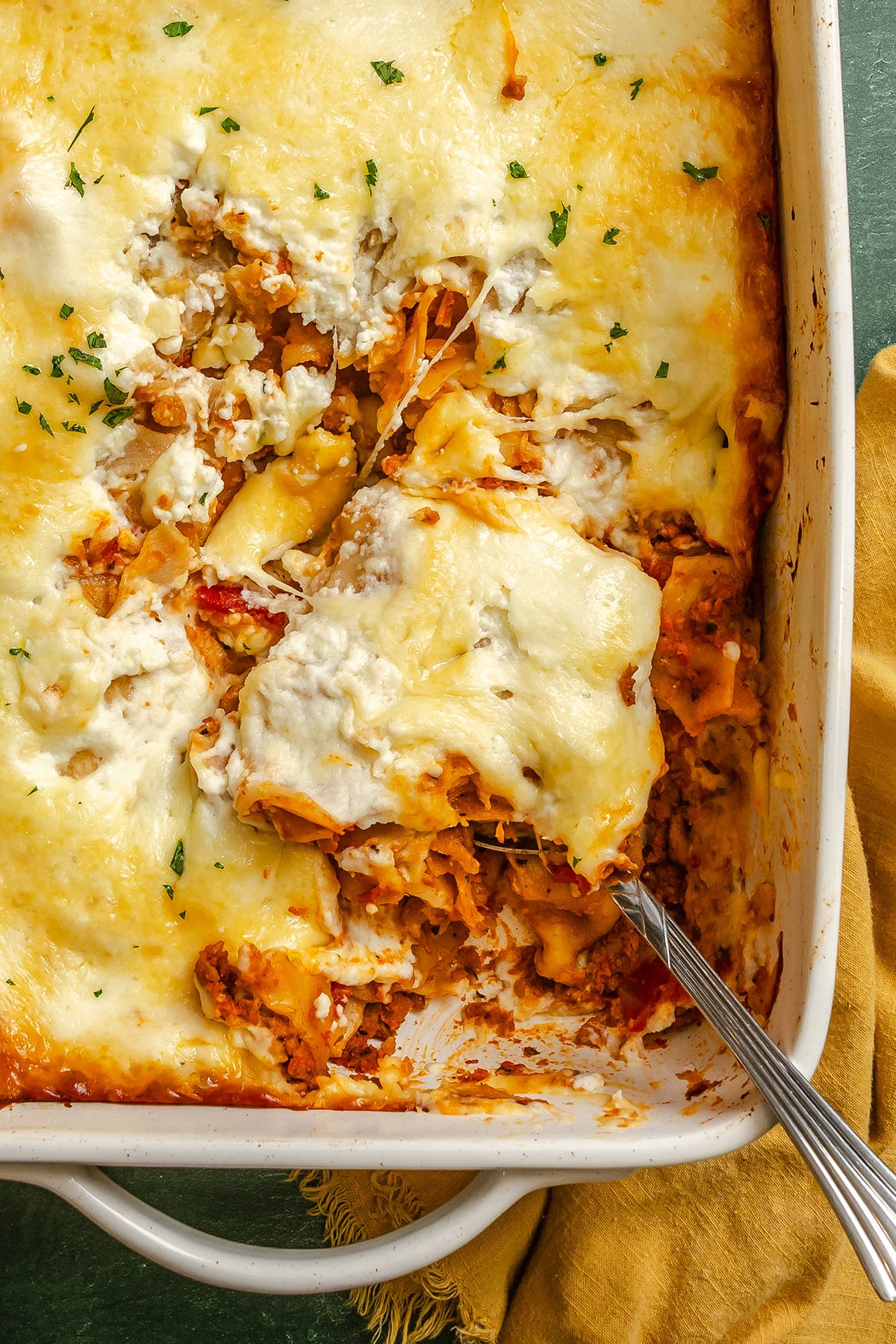 A baked lasagna in a white casserole dish with a serving removed, showing layers of pasta, meat sauce, and melted cheese. A serving spoon rests in the dish.