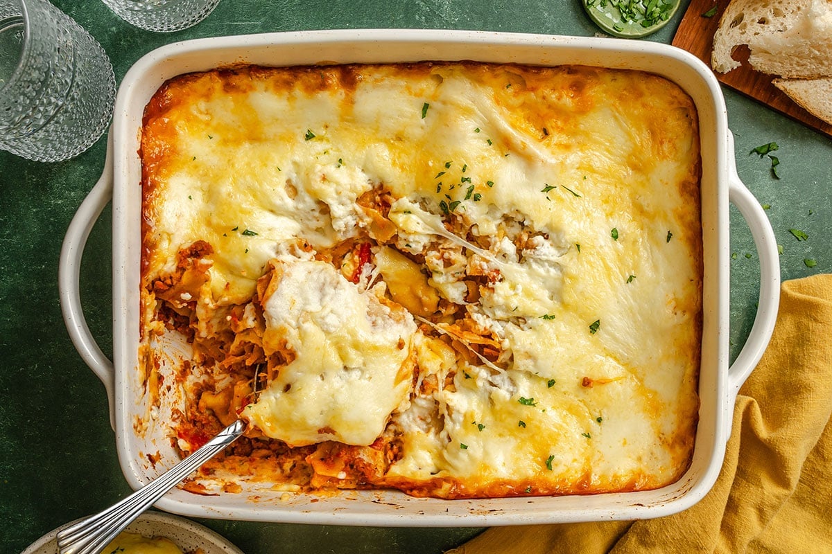 Baked lasagna in a white casserole dish with a serving spoon, topped with melted cheese and sprinkled herbs. Some portions have been served.