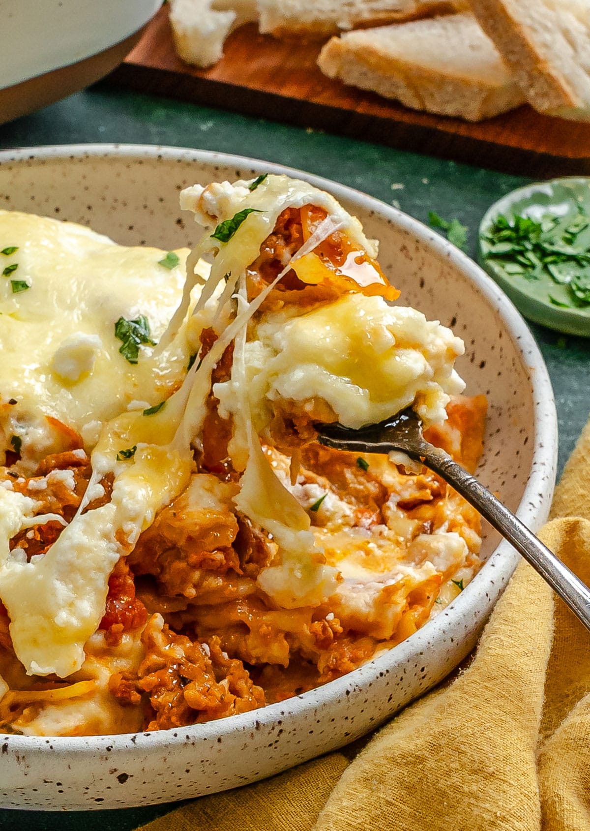 A close-up of a fork lifting a cheesy portion of baked lasagna from a bowl, showing melted cheese, meat sauce, and pasta layers. Slices of bread are in the background.