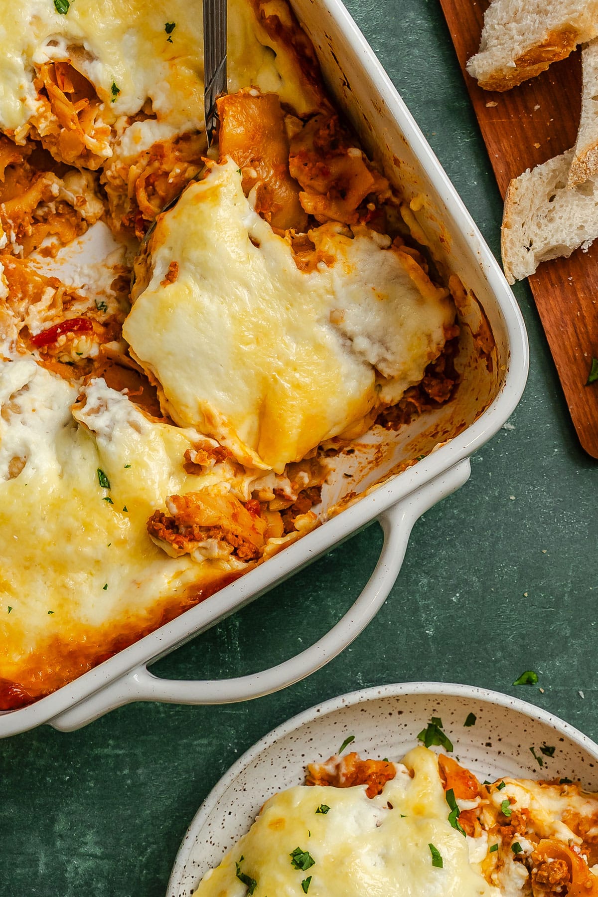 A serving of cheesy baked lasagna is being lifted from a casserole dish, with slices of bread and a plated portion nearby on a green surface.