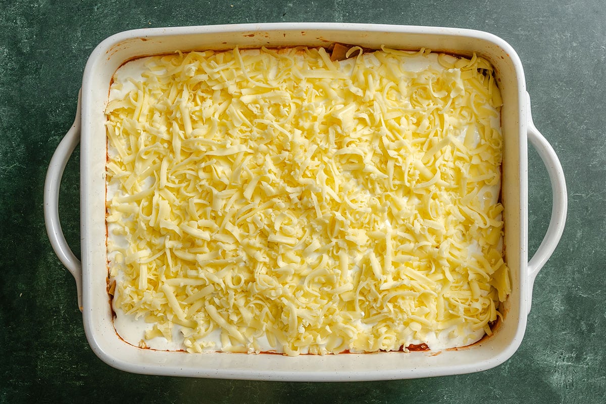 Rectangular baking dish filled with an unbaked casserole topped with a layer of shredded cheese on a dark green surface.