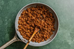 Cooked ground vegan meat with spices in a frying pan, stirred with a wooden spoon, on a green textured surface.