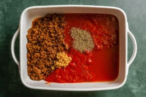 White baking dish containing cooked ground vegan meat, tomato sauce, minced garlic, and a pile of dried Italian herbs, all unmixed, on a dark green surface.