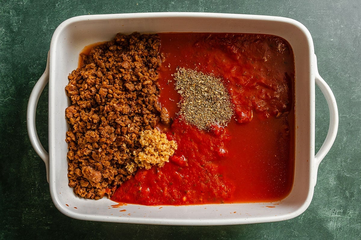 White baking dish containing cooked ground vegan meat, tomato sauce, minced garlic, and a pile of dried Italian herbs, all unmixed, on a dark green surface.