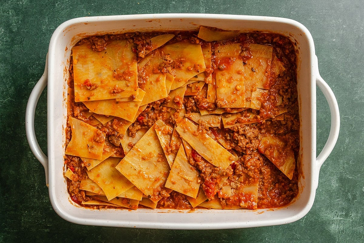 A casserole dish filled with layered pasta sheets and meat sauce, resembling an unbaked or deconstructed lasagna, on a green surface.
