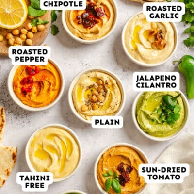 Top-down view of eight bowls of hummus in different flavors, each labeled: chipotle, roasted garlic, roasted pepper, jalapeno cilantro, plain, tahini free, sun-dried tomato, and spinach.