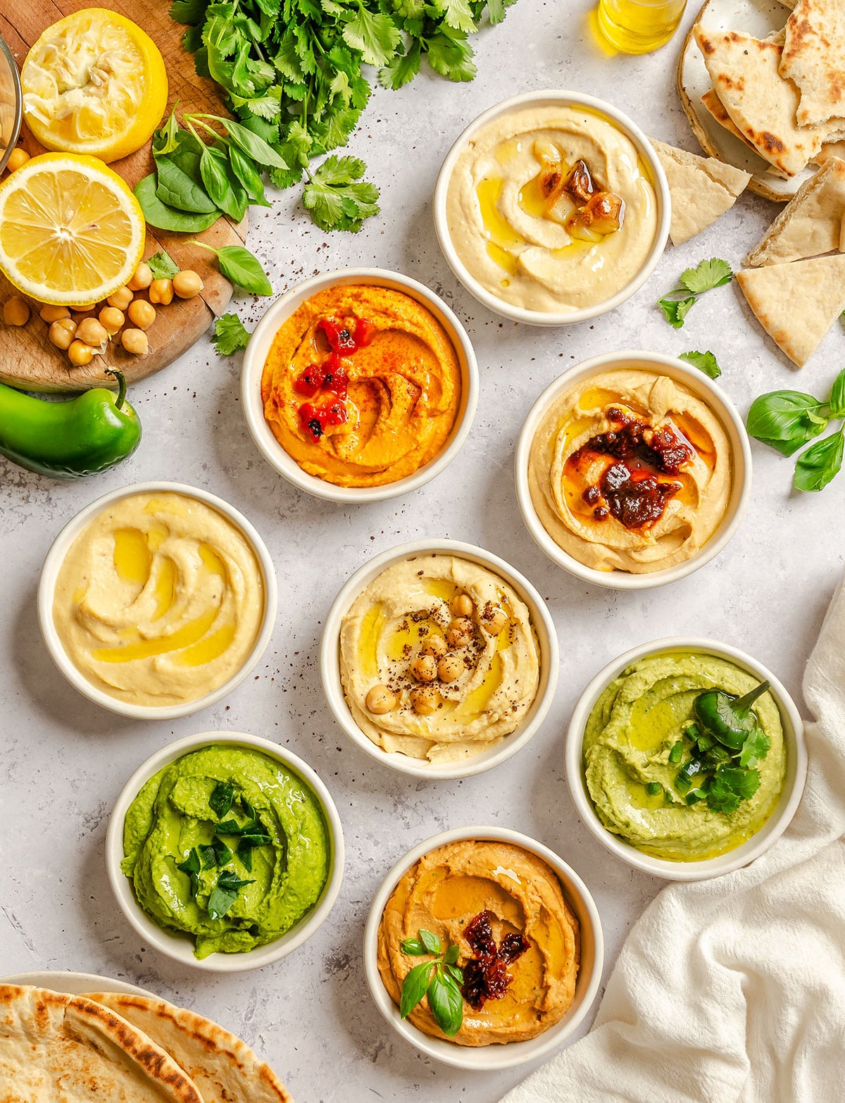 A variety of colorful hummus dips in small bowls, garnished with herbs and spices, surrounded by pita bread, lemon, chickpeas, and fresh greens on a light surface.