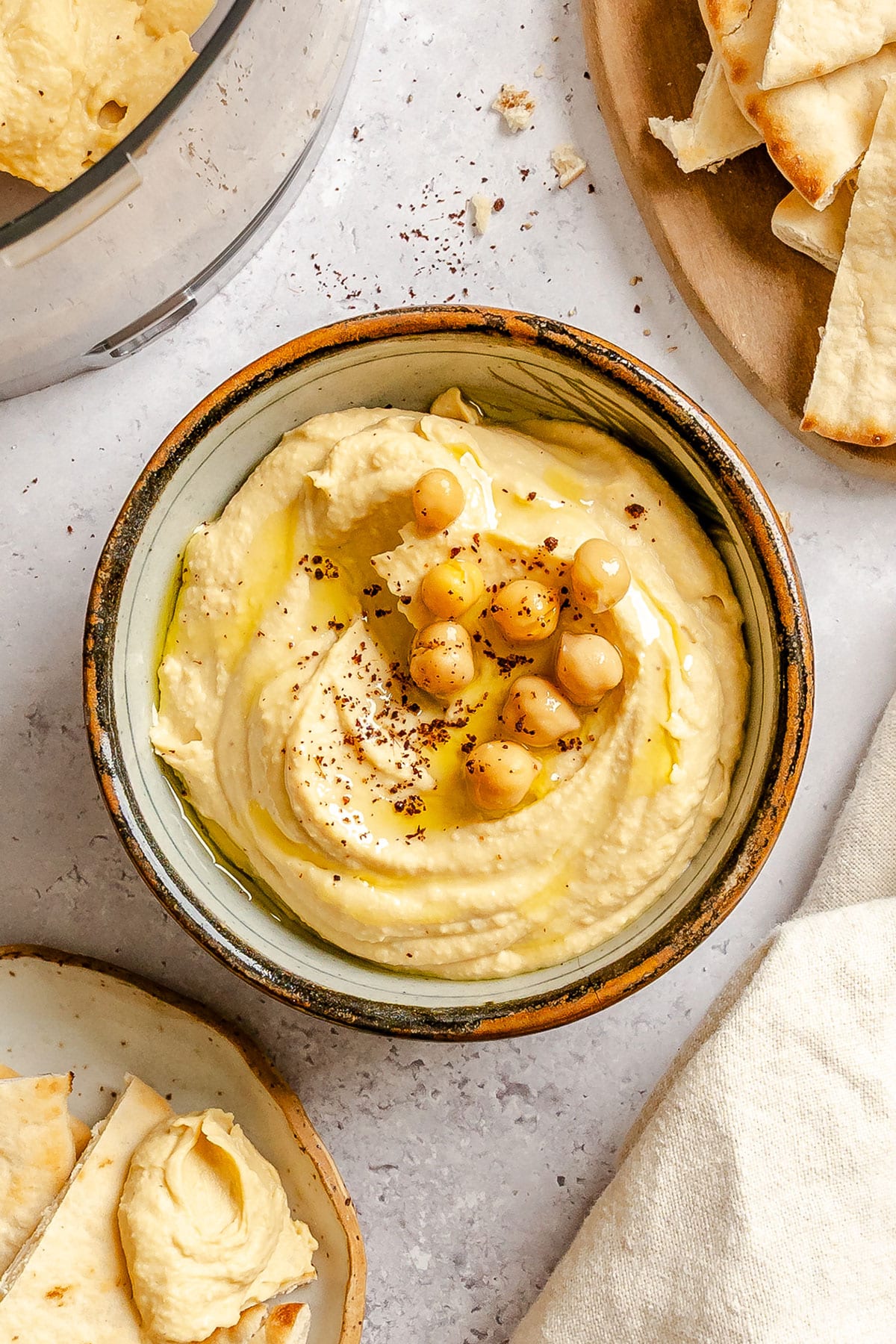 A bowl of hummus topped with whole chickpeas, olive oil, and spices, surrounded by pieces of pita bread on a light surface.