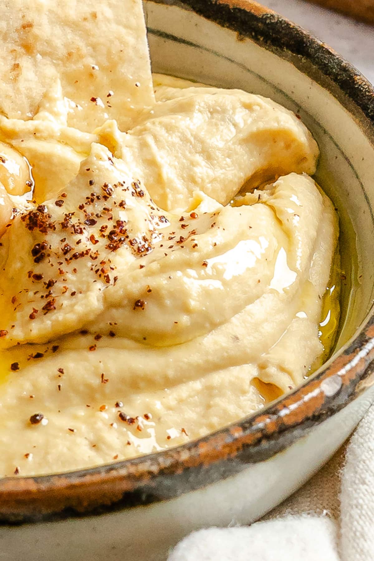 A close-up of creamy hummus in a ceramic bowl, topped with olive oil and spices, with a piece of pita bread dipped in.