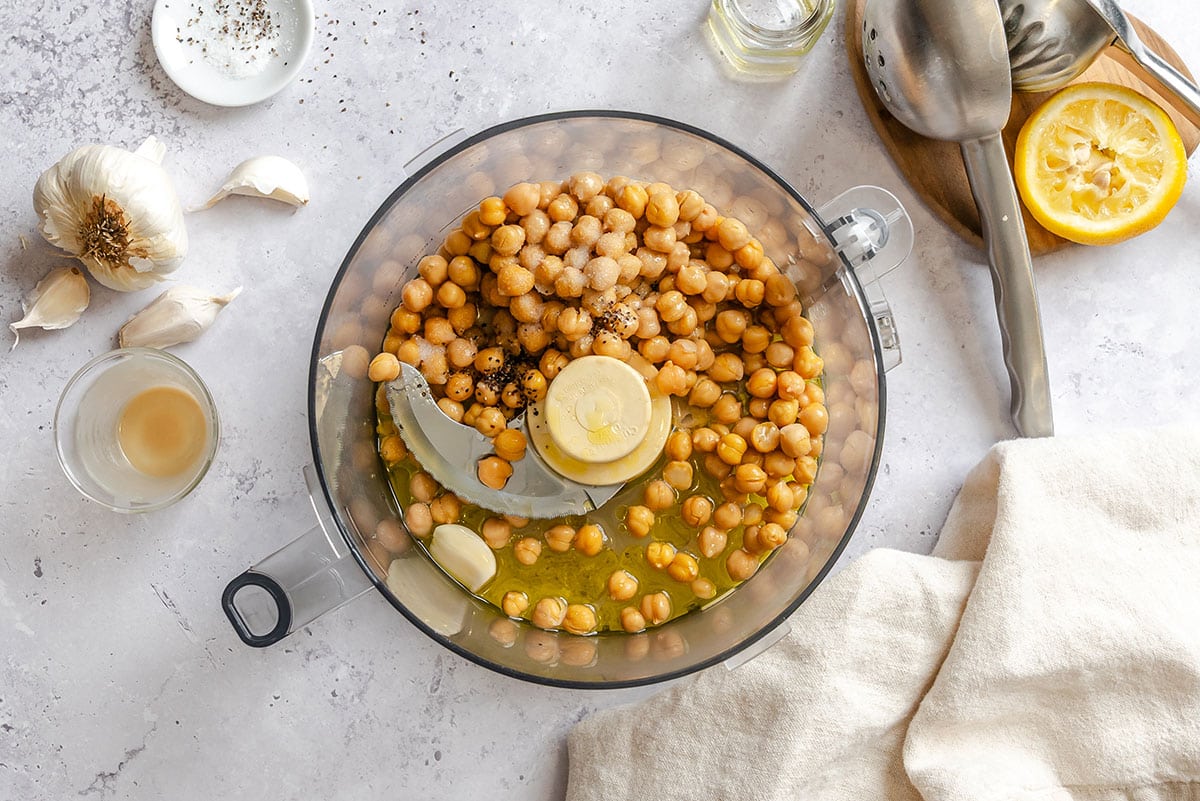 Chickpeas, garlic, lemon juice, and oil in a food processor ready to be blended, surrounded by kitchen utensils and ingredients on a light countertop.