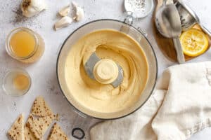 Food processor with smooth hummus, surrounded by flatbread pieces, lemon, garlic cloves, metal utensils, a towel, and liquids in glass jars on a light countertop.