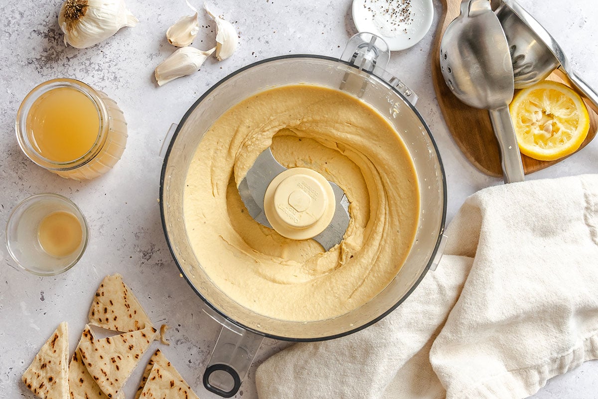 Food processor with smooth hummus, surrounded by flatbread pieces, lemon, garlic cloves, metal utensils, a towel, and liquids in glass jars on a light countertop.