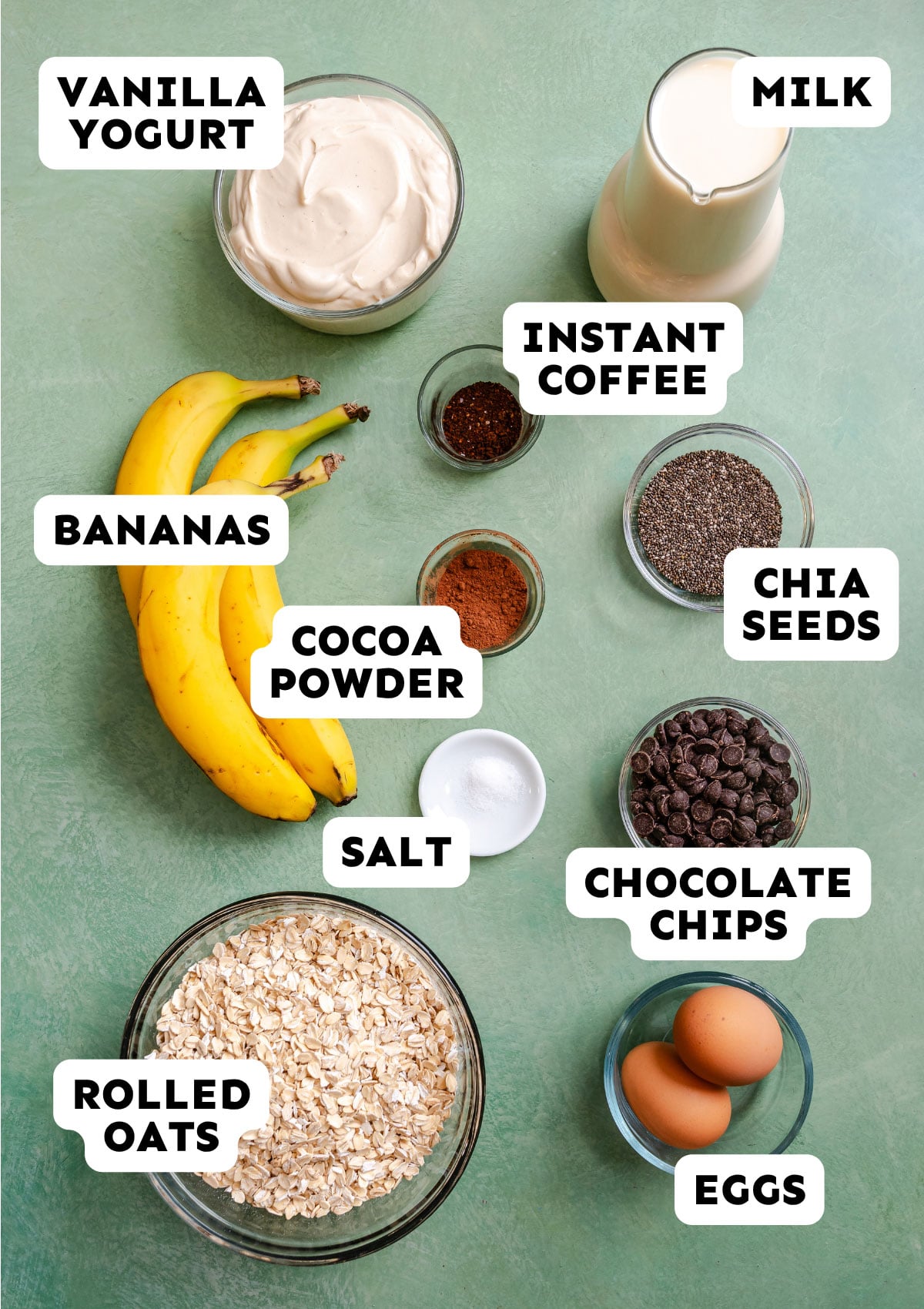 A top-down view of labeled ingredients on a green surface—perfect for Baked Oatmeal or a Dump and Bake Tiramisu—including vanilla yogurt, milk, bananas, instant coffee, cocoa powder, chia seeds, salt, chocolate chips, rolled oats, and eggs.