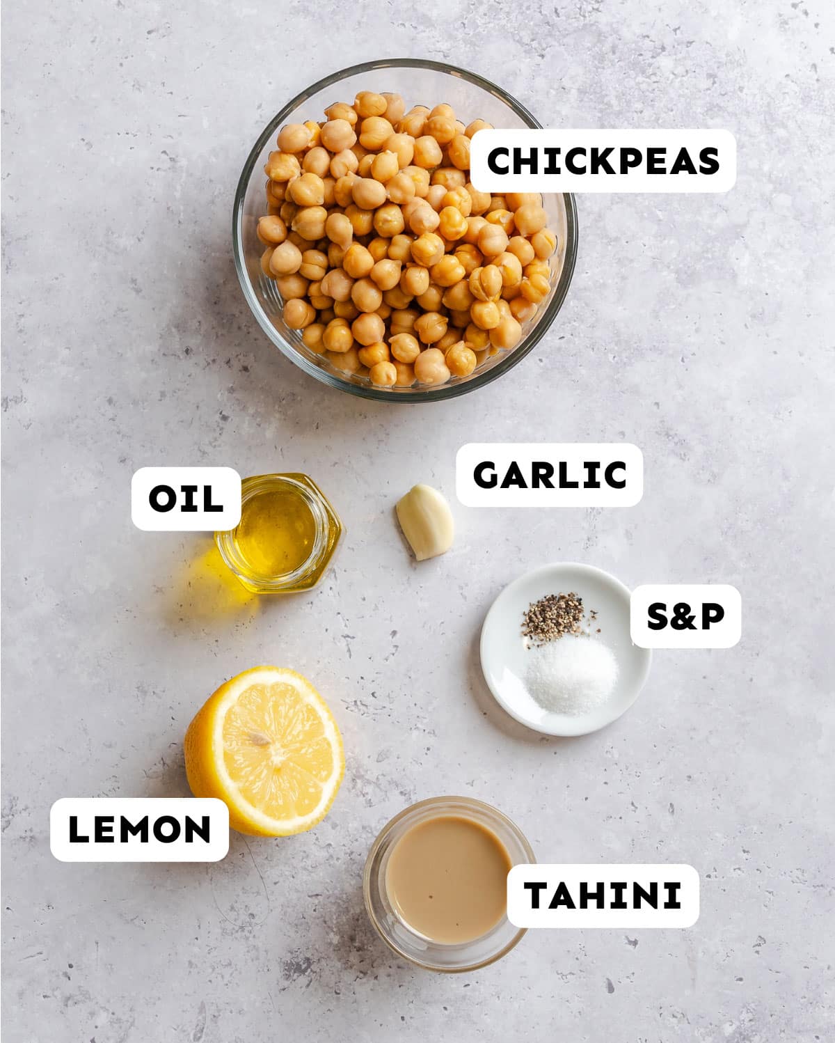 A bowl of chickpeas, a garlic clove, a small glass of oil, half a lemon, a dish of salt and pepper, and a small bowl of tahini arranged on a light surface. Labels identify each ingredient.
