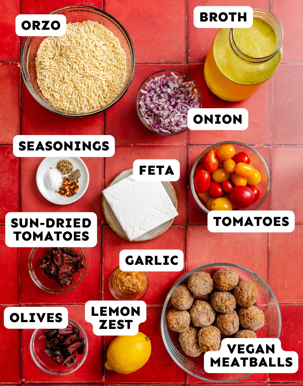 Bowls containing orzo, seasonings, sun-dried tomatoes, olives, lemon zest, feta, garlic, vegan meatballs, cherry tomatoes, chopped onion, and broth on a red tile background.