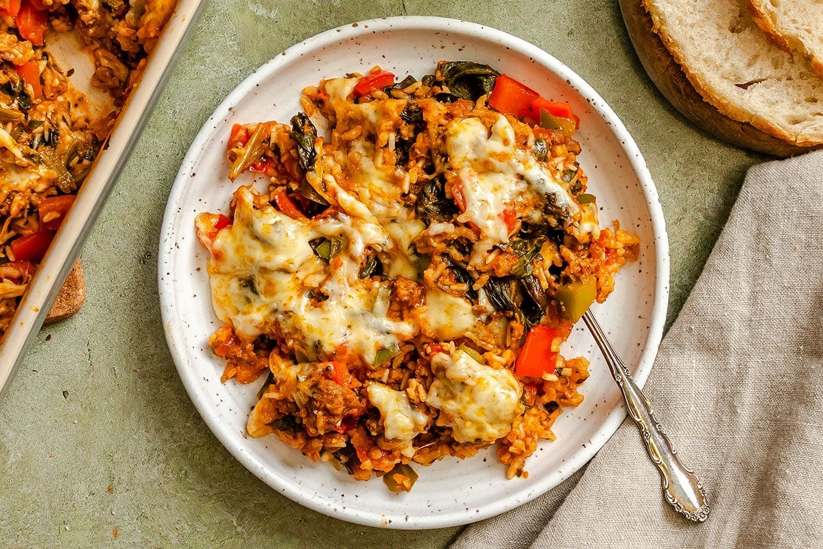 A plate of cheesy baked rice with vegetables and greens, served with a fork, next to slices of bread and a beige napkin.
