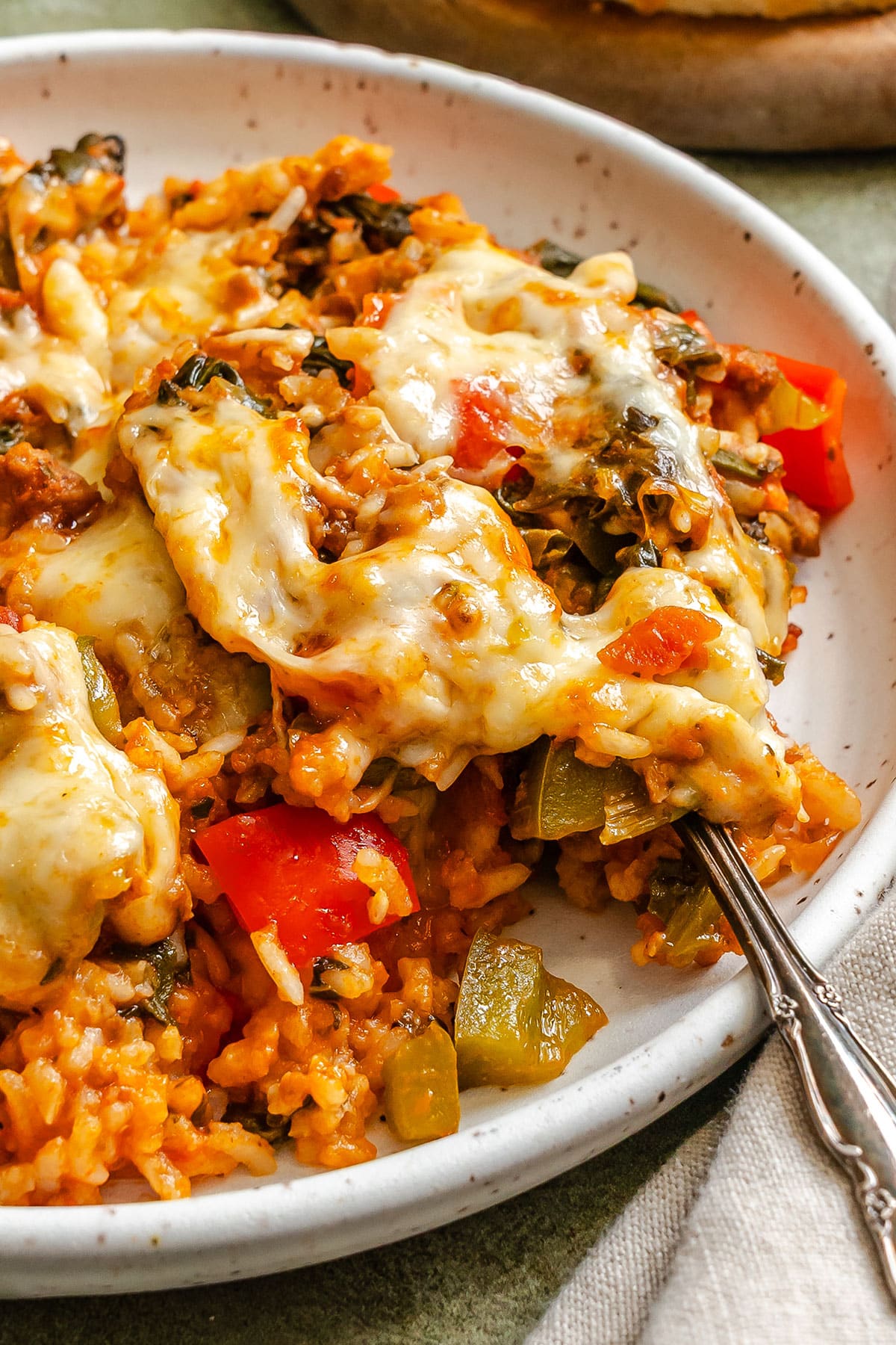 A close-up of a plate of baked rice casserole with melted cheese, red and green bell peppers, and leafy greens, with a fork scooping a portion.