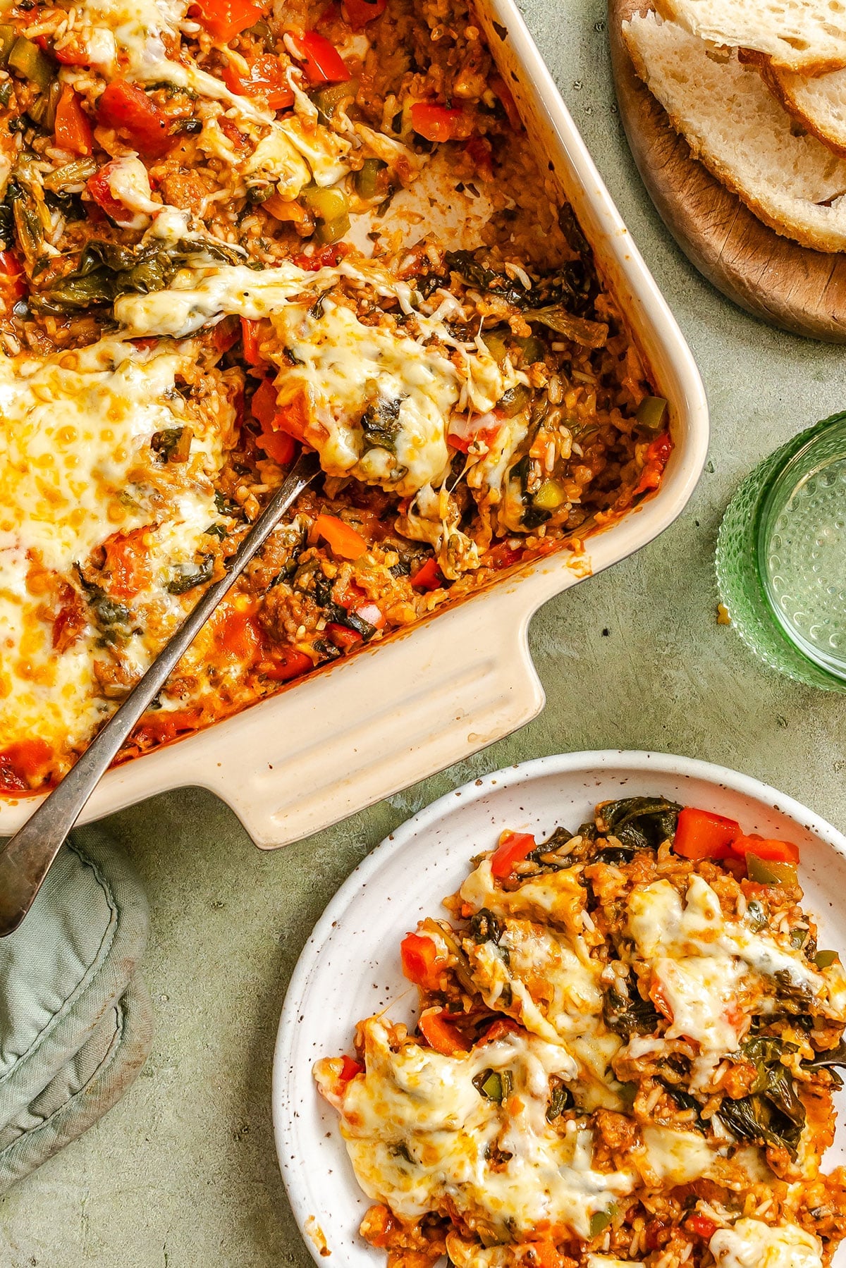 A baked pepper casserole with melted cheese, rice, vegetables, and greens is served in a dish and on a white plate, with a glass of water and sliced bread nearby.