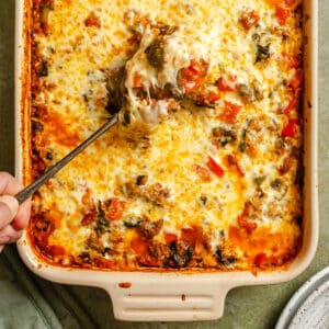 A hand scoops a serving of cheesy baked pepper casserole with vegetables and ground meat from a ceramic dish.