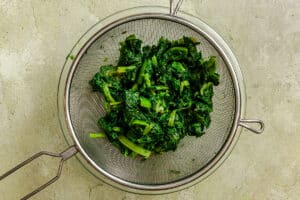Chopped cooked spinach draining in a metal mesh strainer on a light-colored countertop.