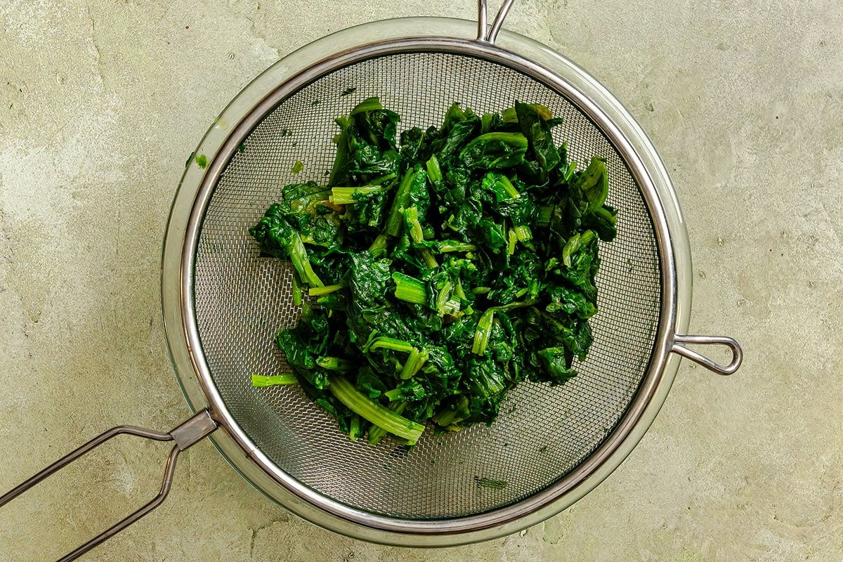 Chopped cooked spinach draining in a metal mesh strainer on a light-colored countertop.