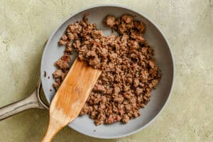 Cooked ground beef in a frying pan with a wooden spatula on a light-colored surface.