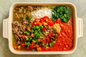 Rectangular casserole dish filled with separated ingredients: cooked ground meat, rice, wilted spinach, diced green and red bell peppers, minced garlic, seasonings, and tomato sauce.