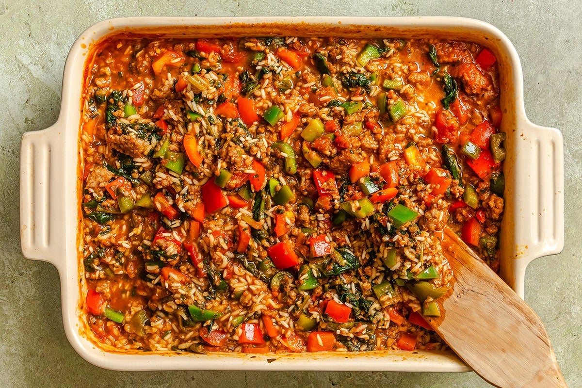 A casserole dish filled with a baked mixture of rice, ground meat, chopped bell peppers, spinach, and tomato sauce, with a wooden spatula resting inside.